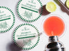 Custom round pulpboard coasters are shown sitting on a white marble surface next to lemons and an azalea cocktail being poured from a silver cocktail shaker. The custom coasters feature a golf club lounge design with an illustration of a golf ball on a tee and people cheersing drinks together along with a striped border. The established year, name, lounge name, and tagline can be customized.