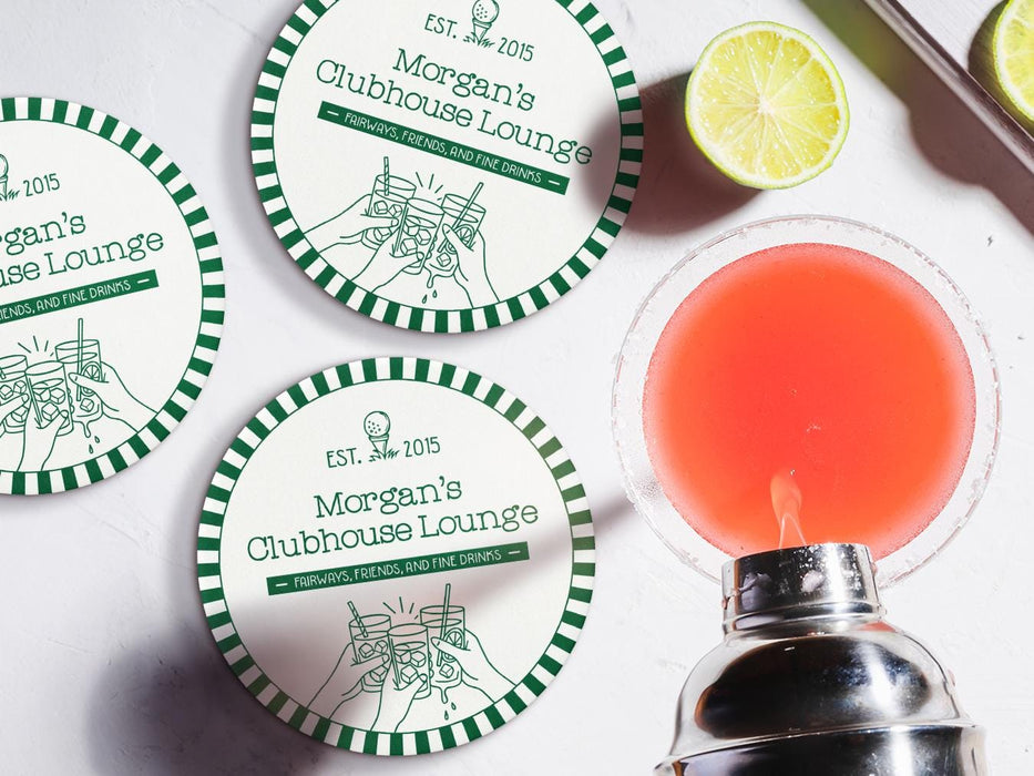 Custom round pulpboard coasters are shown sitting on a white marble surface next to lemons and an azalea cocktail being poured from a silver cocktail shaker. The custom coasters feature a golf club lounge design with an illustration of a golf ball on a tee and people cheersing drinks together along with a striped border. The established year, name, lounge name, and tagline can be customized.