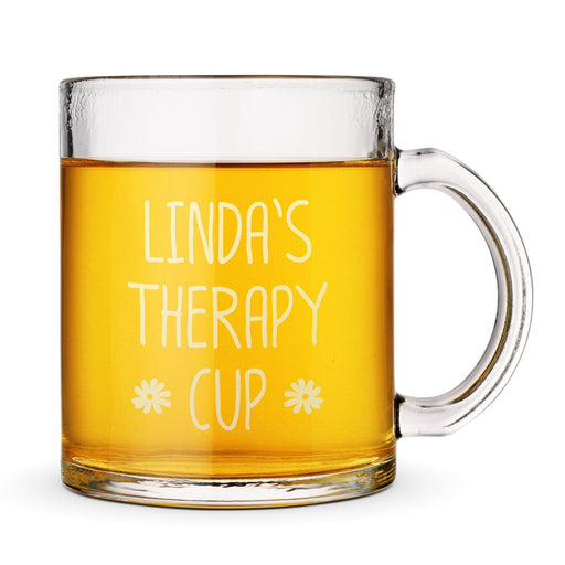 A clear glass mug filled with a yellow liquid, likely tea or coffee, with the text "Linda's Therapy Cup" engraved on it.