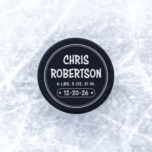 Personalized hockey puck with a white UV printed round birth announcement design displayed on an icy surface.