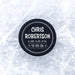 Personalized hockey puck with a white UV printed round birth announcement design displayed on an icy surface.
