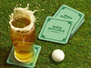 Custom square pulpboard coasters are shown sitting on a grassy, green golf course next to a pint of beer and a white golf ball. The pint of beer has a golf ball splashing into it and has a coaster underneath it. The custom coasters feature a green golf bar design with interlocking golf clubs used as a border. The bar name and location can be customized.