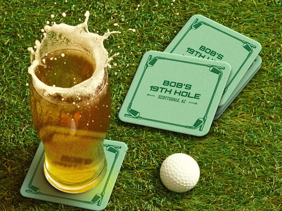 Custom square pulpboard coasters are shown sitting on a grassy, green golf course next to a pint of beer and a white golf ball. The pint of beer has a golf ball splashing into it and has a coaster underneath it. The custom coasters feature a green golf bar design with interlocking golf clubs used as a border. The bar name and location can be customized.
