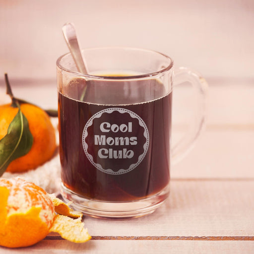 A glass mug filled with a dark liquid, likely coffee, sits on a wooden surface. The mug has an engraving graphic that reads "Cool Moms Club". Next to the mug are a peeled orange and a spoon.