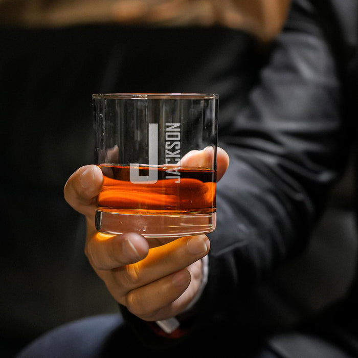 Close-up lifestyle photo of a personalized whiskey glass held in hand, showing the large initial and vertical name engraving. The glass is filled with whiskey and ice, ideal for gifting.