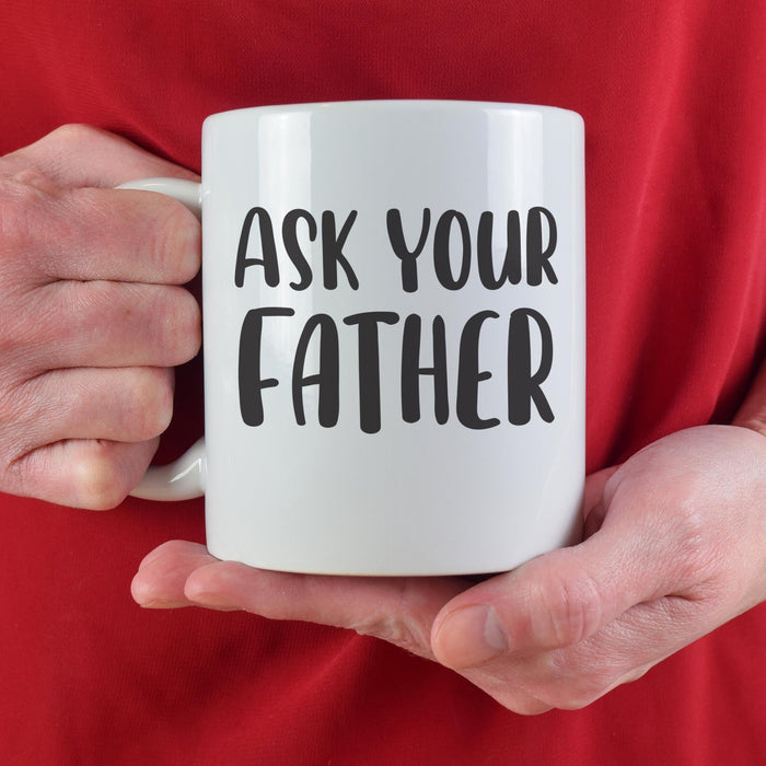 A person in a red sweater holding the “Ask Your Father” mug with both hands. The bright cozy setting adds a fun and playful gift feel.