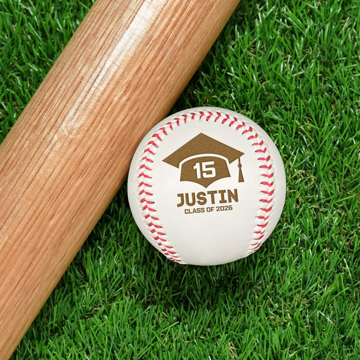 A baseball bat and a baseball with a personalized graduation cap-themed design, rests on a grassy surface.