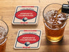 Custom square pulpboard coasters are shown sitting on a wooden table next to two drink glasses. The custom coasters feature a 19th hole bar design with an illustration of a golf ball. The tagline, bar name, established year, and location can be customized on these.