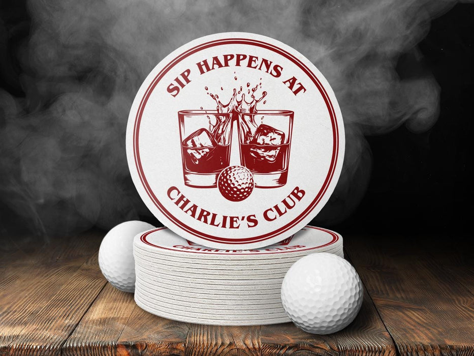 A stack of custom pulpboard coasters and two white golf balls are shown in front of a black, smoky background while sitting on a dark wooden table surface. The custom coasters feature a golf ball centered between two whiskey glasses clinking together and splashing out with customizable text wrapping around the illustration. The coaster design is printed in ruby red.