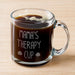 A glass mug filled with a dark liquid, likely coffee, with foam on top. The mug has a handle and is placed on a wooden surface.