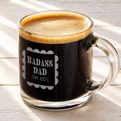 A mug of dark coffee with a foam top, sitting on a wooden surface. It has the engraved design on it that reads "Badass Dad From, Russel".