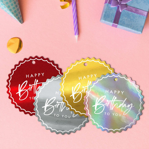 Four elegant script hang tags in metallic gold, metallic silver, glitter red, and glitter rainbow lying beside colorful gifts and party decorations.