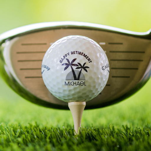 Close-up of a custom retirement golf ball featuring palm trees and a personalized name. Displayed on a tee in front of a golf club.