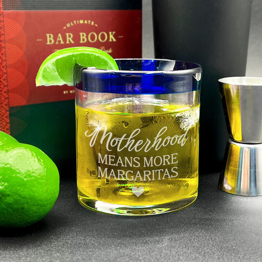 A glass of yellow liquid with a lime wedge, a book, and a silver shaker on a table. The engraving on the glass reads "Motherhood means more margaritas"