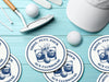 Custom round pulpboard coasters are shown sitting on a bright blue wooden surface. Golf balls, white golf tees, a golf club, and golf hat are shown next to the coasters. The custom coasters feature a golf ball centered between two whiskey glasses clinking together and splashing out with customizable text wrapping around the illustration. The coaster design is printed in denim.