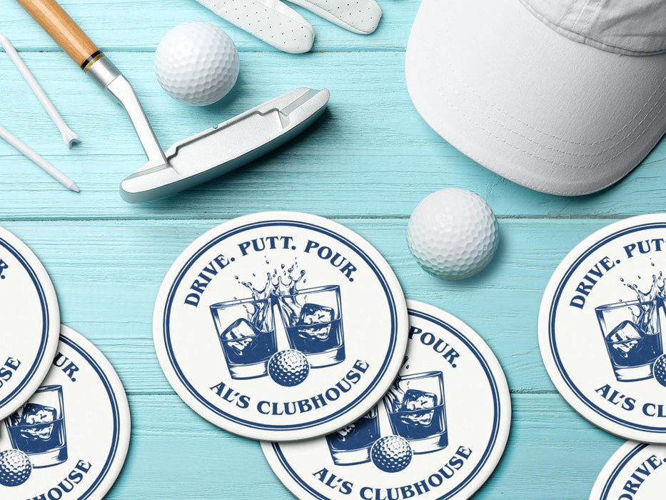 Custom round pulpboard coasters are shown sitting on a bright blue wooden surface. Golf balls, white golf tees, a golf club, and golf hat are shown next to the coasters. The custom coasters feature a golf ball centered between two whiskey glasses clinking together and splashing out with customizable text wrapping around the illustration. The coaster design is printed in denim.