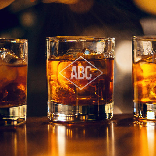 Warm bar-style lifestyle image of a personalized whiskey glass with glowing ambient lights in the background, featuring a diamond-shaped “ABC” initials engraving. The lighting highlights the clean and modern monogram design.