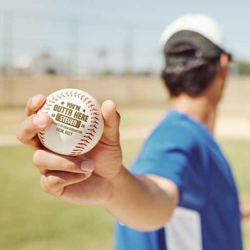 A person is holding a personalized baseball in their hand, with a blurred background that appears to be a baseball field.
