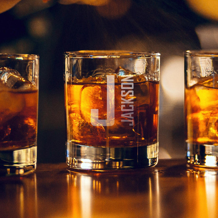 Warm bar-style lifestyle image with glowing ambient lights in the background. The personalized glass is filled with whiskey and ice, highlighting the bold custom engraving.