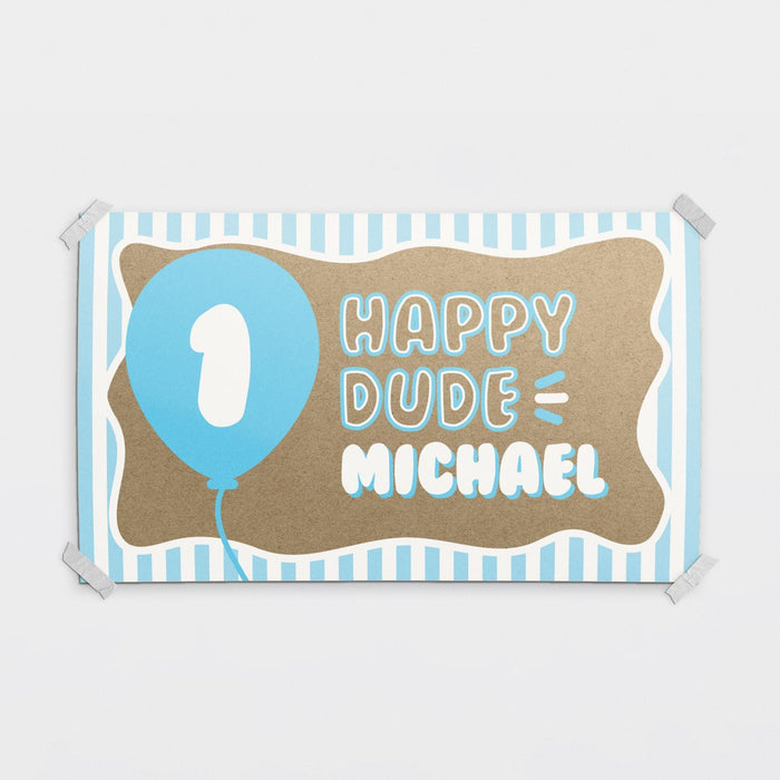 Personalized ONE Happy Dude first birthday banner printed on brown butcher paper with blue balloon graphic, striped border details, and custom name personalization.
