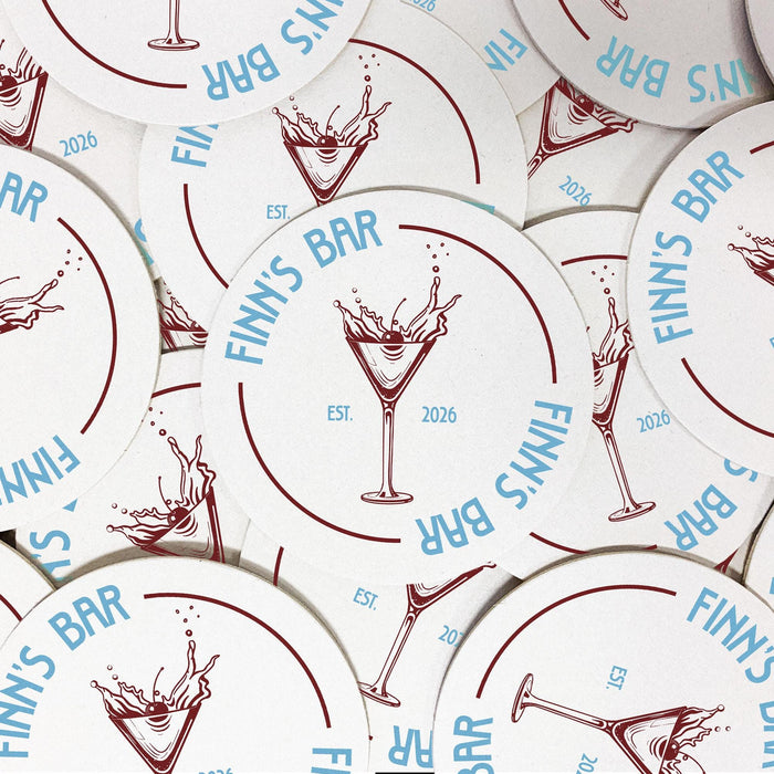 Several personalized martini coasters spread across a tabletop, each featuring the custom bar name, martini glass illustration, and established year.