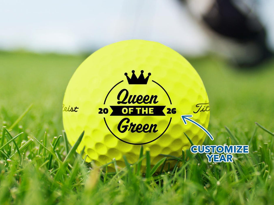 Custom yellow Titleist golf ball shown with a Queen of the Green design printed on it. The golf ball sits on a grassy golf course.