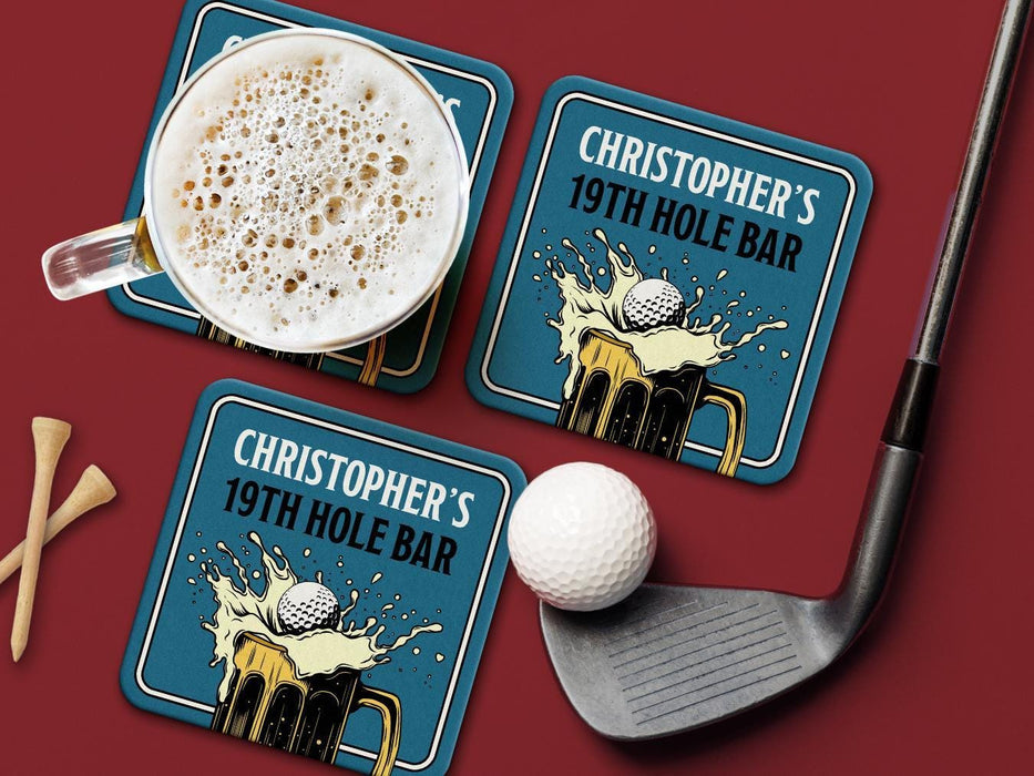 Custom square pulpboard coasters are shown sitting on a dark red surface. Golf tees, a golf ball, and a golf club sit beside them. One coaster has a beer mug on top of it. The custom coasters feature a 19th hole bar design with an illustration of a golf ball splashing into a beer mug and a blue background.