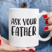 Close-up of hands holding the white “Ask Your Father” ceramic mug with denim clothing in the background. A casual lifestyle image emphasizing the mug’s everyday use and gift appeal.