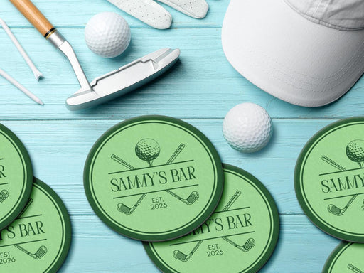 Custom round pulpboard coasters are shown sitting on a bright blue wooden surface. Golf balls, white golf tees, a golf club, and golf hat are shown next to the coasters. The custom coasters feature a green, elegant golf bar design with interlocking golf clubs and a golf ball sitting on top of a golf tee. The bar name and established year can be customized.