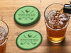 Custom round pulpboard coasters are shown sitting on a wooden table next to two drink glasses. The custom coasters feature a green, elegant golf bar design with interlocking golf clubs and a golf ball sitting on top of a golf tee. The bar name and established year can be customized.