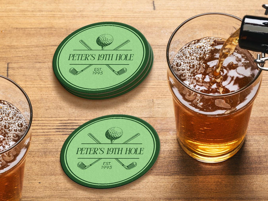 Custom round pulpboard coasters are shown sitting on a wooden table next to two drink glasses. The custom coasters feature a green, elegant golf bar design with interlocking golf clubs and a golf ball sitting on top of a golf tee. The bar name and established year can be customized.