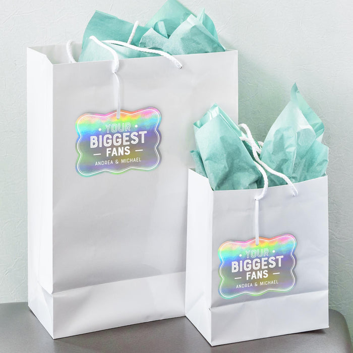 White gift bags with tissue paper decorated using personalized “Your Biggest Fans” wedding gift tags in holographic rainbow foil. A stylish way to add a custom touch to wedding favors and gift packaging.