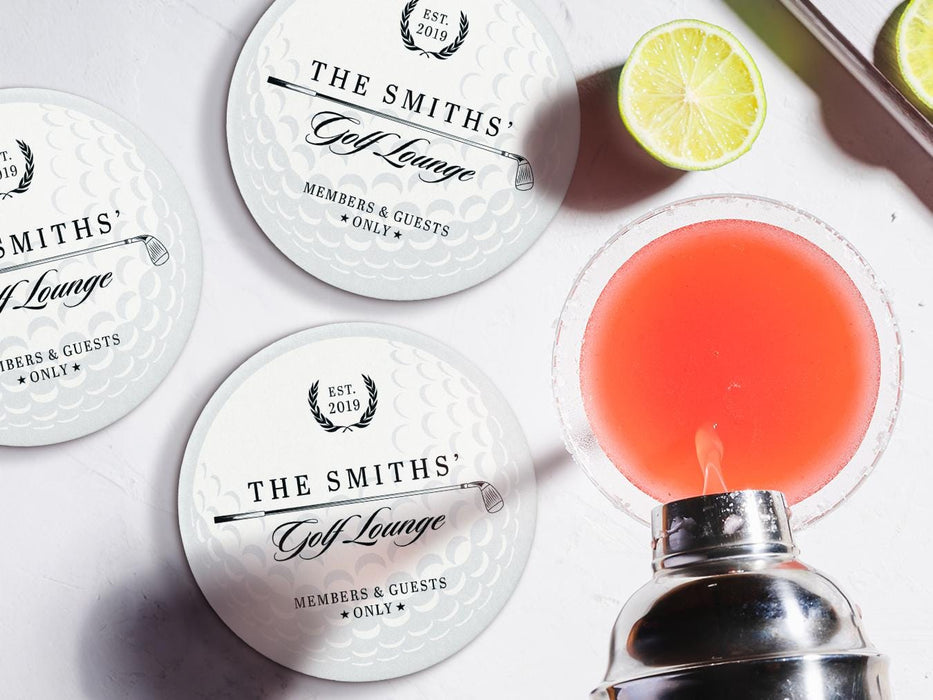 Custom round pulpboard coasters are shown sitting on a white marble surface next to lemons and an azalea cocktail being poured from a silver cocktail shaker. The custom coasters feature a golf club and overlayed golf ball design with the text Members & Guests Only between stars. The last name, club name, and established year can be customized.