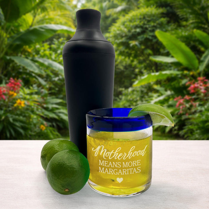 A black cocktail shaker, a glass with a yellow drink and a lime, with a blurred background of greenery.