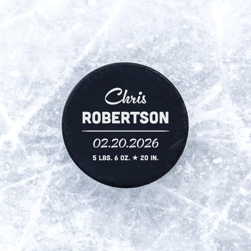 Personalized hockey puck with a white UV printed script name and birth stats announcement design displayed on an icy surface.