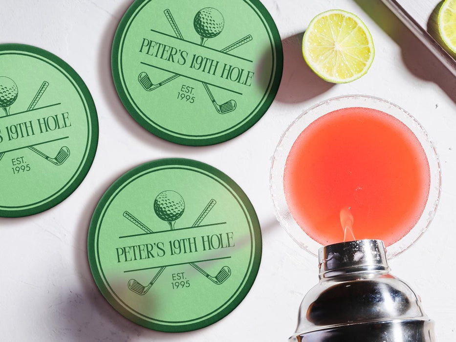 Custom round pulpboard coasters are shown sitting on a white marble surface next to lemons and an azalea cocktail being poured from a silver cocktail shaker. The custom coasters feature a green, elegant golf bar design with interlocking golf clubs and a golf ball sitting on top of a golf tee. The bar name and established year can be customized.