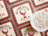 Custom square pulpboard coasters are shown laid out on a wooden surface. A beer mug is shown on top of a few of the coasters. The coasters feature design with an illustration of a golf ball on a tee, checkerboard border, and custom text.