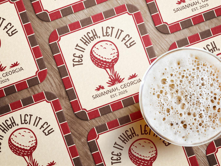 Custom square pulpboard coasters are shown laid out on a wooden surface. A beer mug is shown on top of a few of the coasters. The coasters feature design with an illustration of a golf ball on a tee, checkerboard border, and custom text.