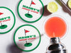 Custom round pulpboard coasters are shown sitting on a white marble surface next to lemons and an azalea cocktail being poured from a silver cocktail shaker. The custom coasters features a green border, golf ball, a red flag, and golf hole illustration and bold text across the center that can be customized with a name and lounge name.