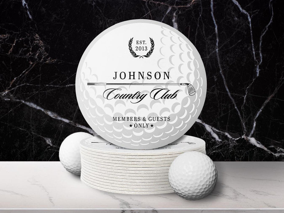 A stack of custom pulpboard coasters and two white golf balls are shown in front of a black marble background while sitting on a white marble surface. The coasters feature a golf club and overlayed golf ball design with the text Members & Guests Only between stars. The last name, club name, and established year can be customized.