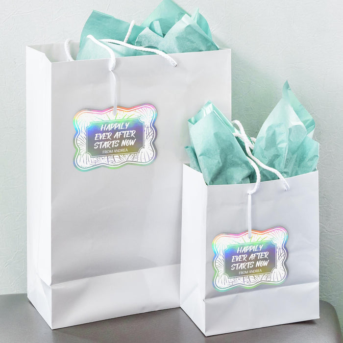 White gift bags with pastel tissue paper featuring personalized rainbow foil wedding gift tags. The custom tags are attached with string and styled for weddings, bridal showers, or event favors.