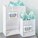 White gift bags with pastel tissue paper featuring personalized rainbow foil wedding gift tags. The custom tags are attached with string and styled for weddings, bridal showers, or event favors.