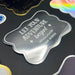 Close-up of a metallic silver “Let Your Adventure Begin” wedding gift tag on black cardstock. The personalized foil cardstock tag features custom sender names with elegant decorative accents.