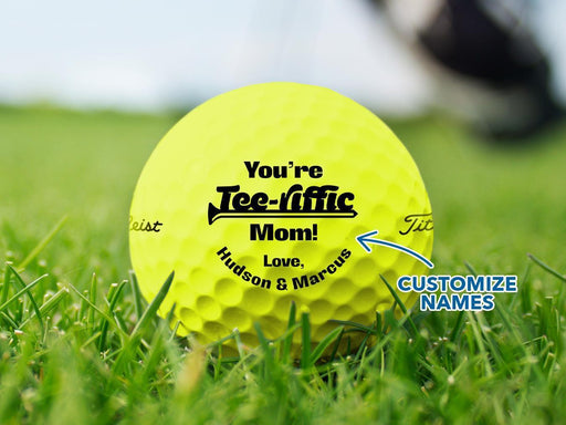 Custom yellow Titleist golf ball shown with a You're Tee-riffic Mom! design printed on it. The golf ball sits on a grassy golf course.