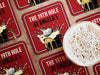 Custom square pulpboard coasters are shown laid out on a wooden surface. A beer mug is shown on top of a few of the coasters. The custom coasters feature a 19th hole bar design with an illustration of a golf ball splashing into a beer mug and a red background.