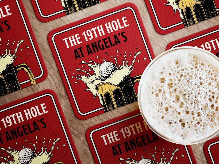 Custom square pulpboard coasters are shown laid out on a wooden surface. A beer mug is shown on top of a few of the coasters. The custom coasters feature a 19th hole bar design with an illustration of a golf ball splashing into a beer mug and a red background.