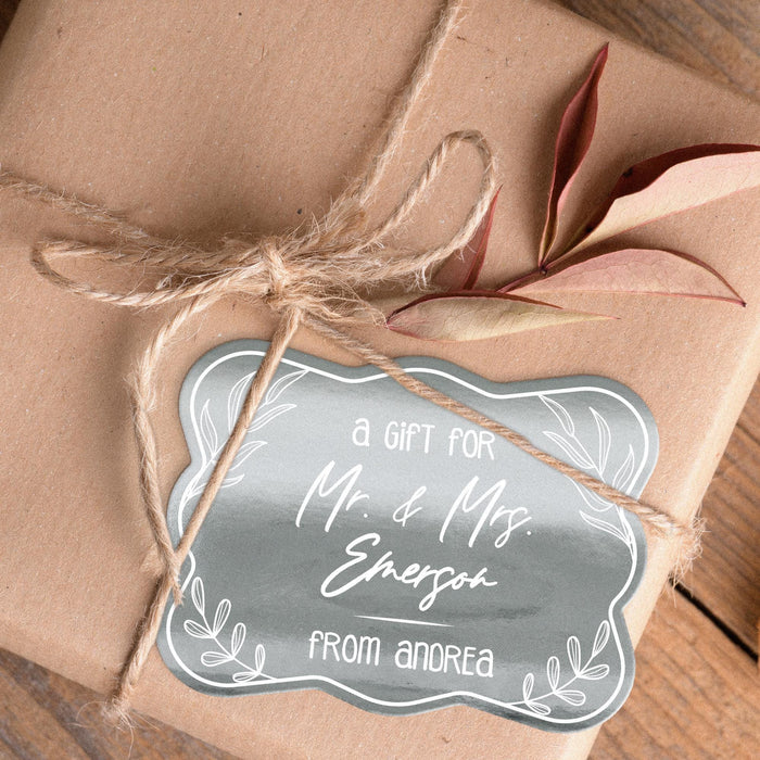 Close-up of a metallic silver “Mr. & Mrs.” wedding gift tag tied onto blush fabric packaging with twine. The foil cardstock tag features custom names in an elegant script font.