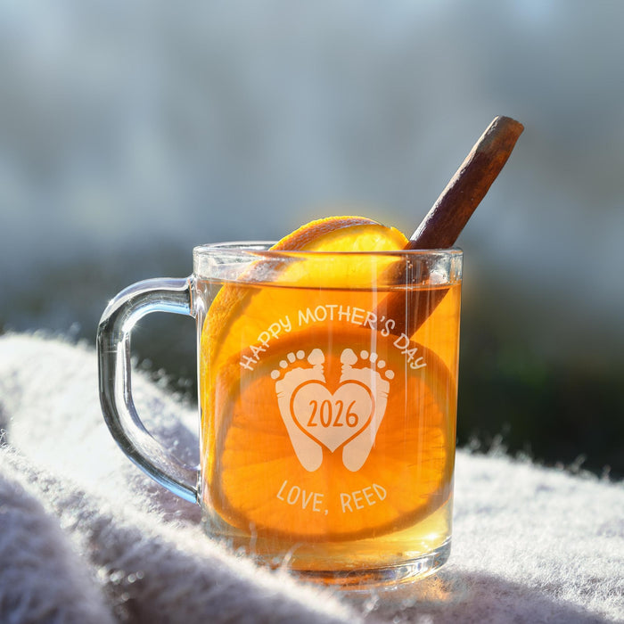A glass mug filled with a warm beverage, likely tea, and garnished with an orange slice and a cinnamon stick. The mug has a personalized message engraved on it, celebrating Mother's Day.