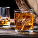 Lifestyle image of the personalized whiskey glass with whiskey and large ice cubes placed on a wooden table beside another drink. The “Brandon’s Lounge” engraving gives the glass a refined and personalized bar-inspired look.