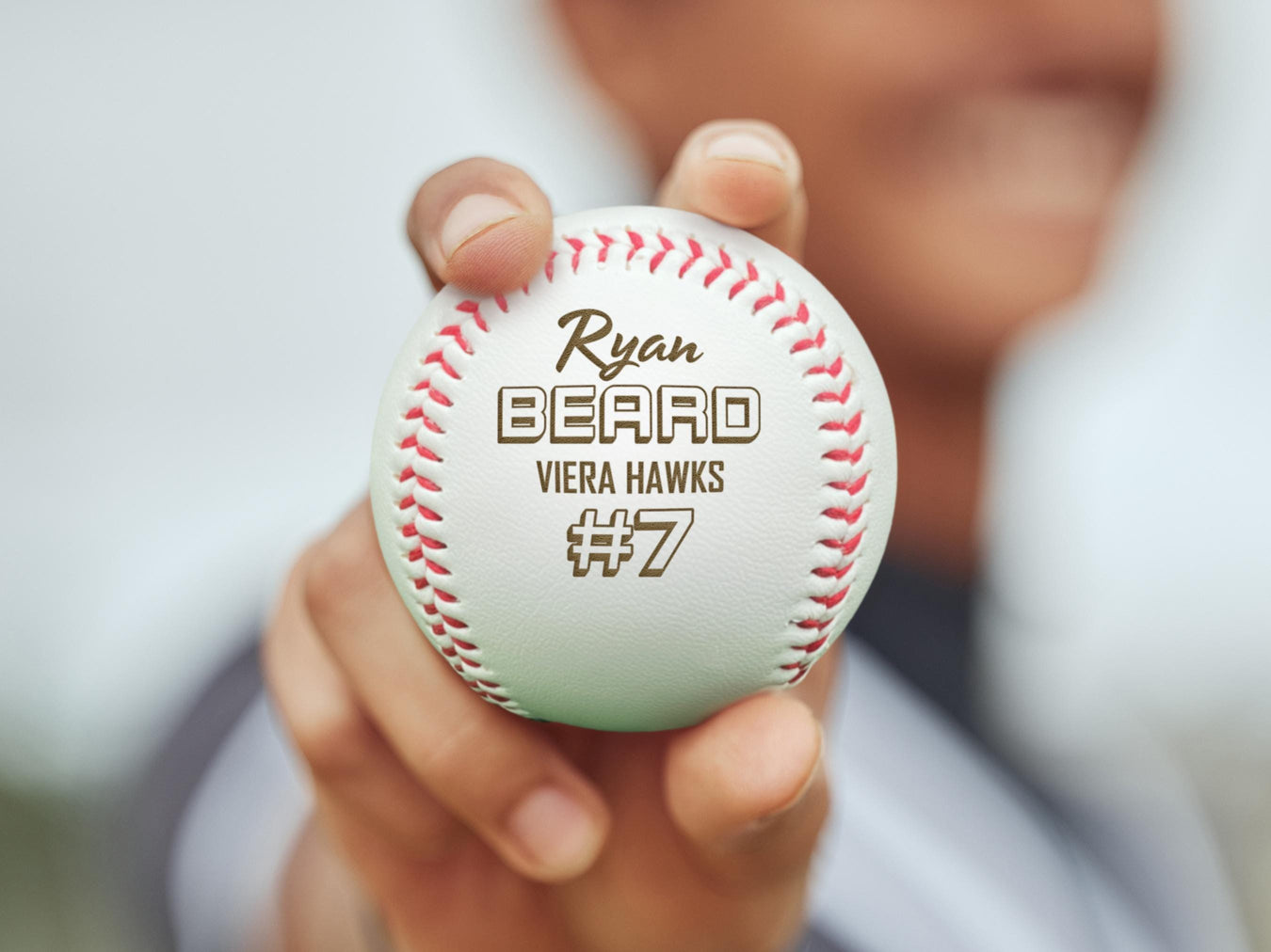 A baseball player holds up an engraved baseball outside. The baseball has the player's name, team name, and number engraved into it. This design features stylized text using script and 3D block lettering.